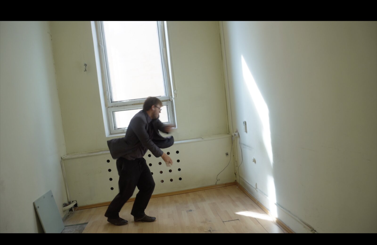 Future Days still — a figure in motion inside a bare room with a perforated wall panel